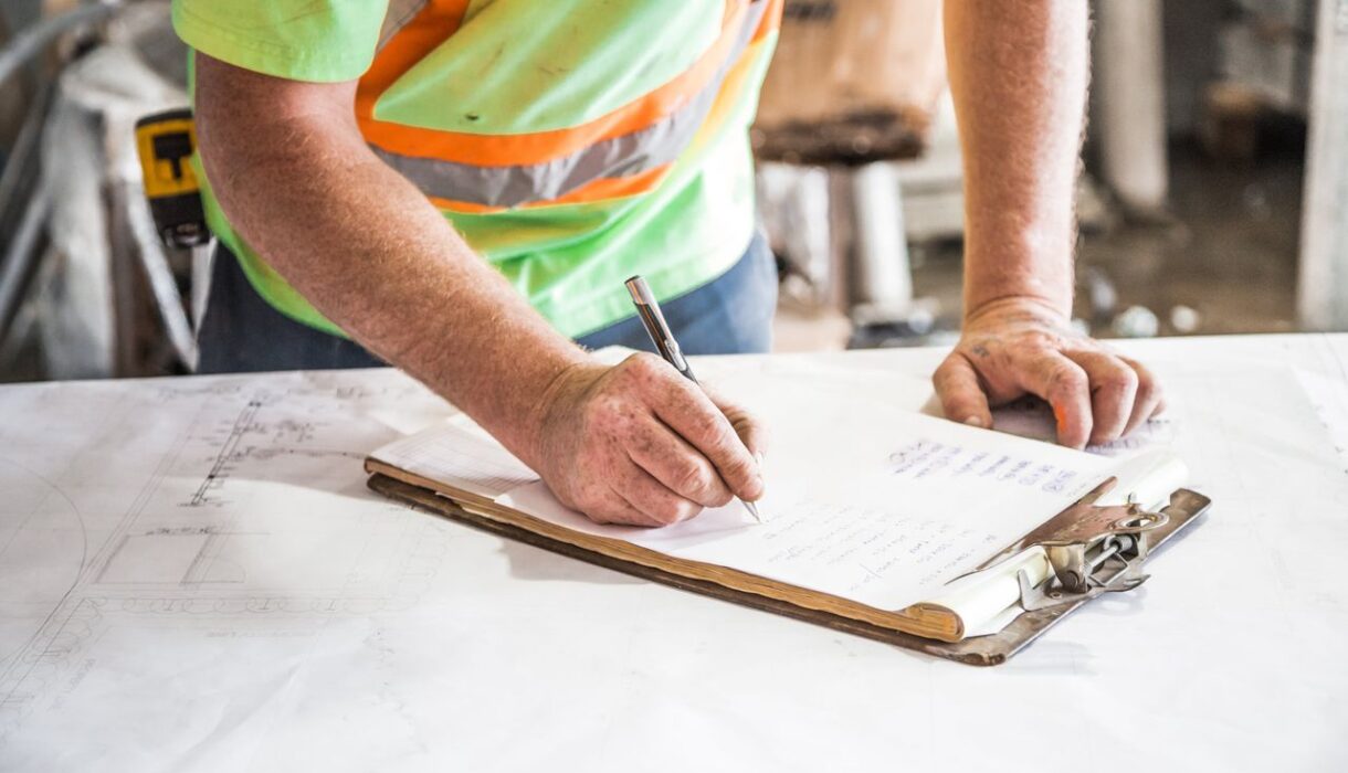 A construction worker writes notes on a clipboard at a construction site, emphasizing planning and precision.