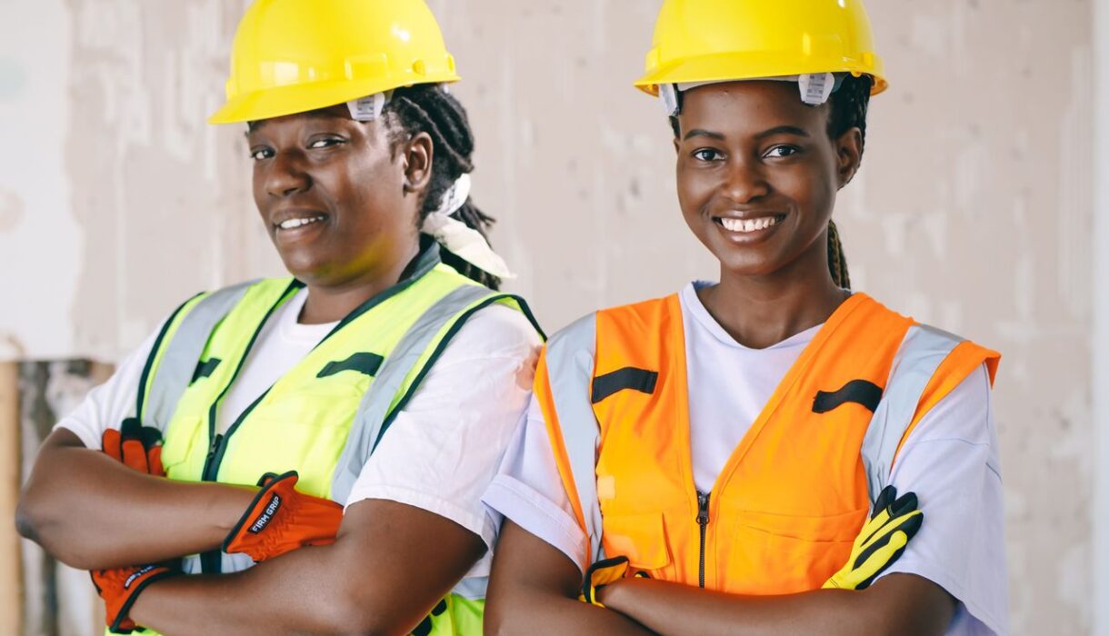 Two female construction workers wearing protective gear and smiling confidently indoors.